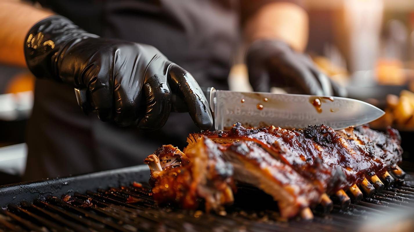 $!Grill restaurant kitchen. Chef in black cooking gloves using knife to cut smoked pork ribs.